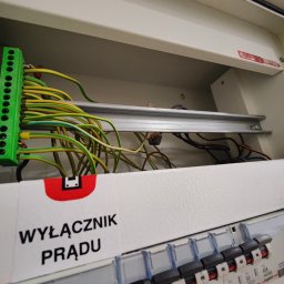 Jarosław Gołembiewski - Otwarta skrzynka elektryczna z podłączonymi przewodami (żółto-zielone) do listwy zaciskowej. Widoczne bezpieczniki i etykieta 'Wyłącznik prądu'.