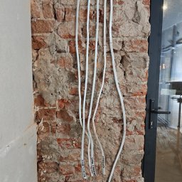 Jarosław Gołembiewski - Białe przewody elektryczne prowadzone po surowej, ceglanej ścianie z widocznymi oznaczeniami, obok ściany otynkowanej i czarnej ramy okiennej.