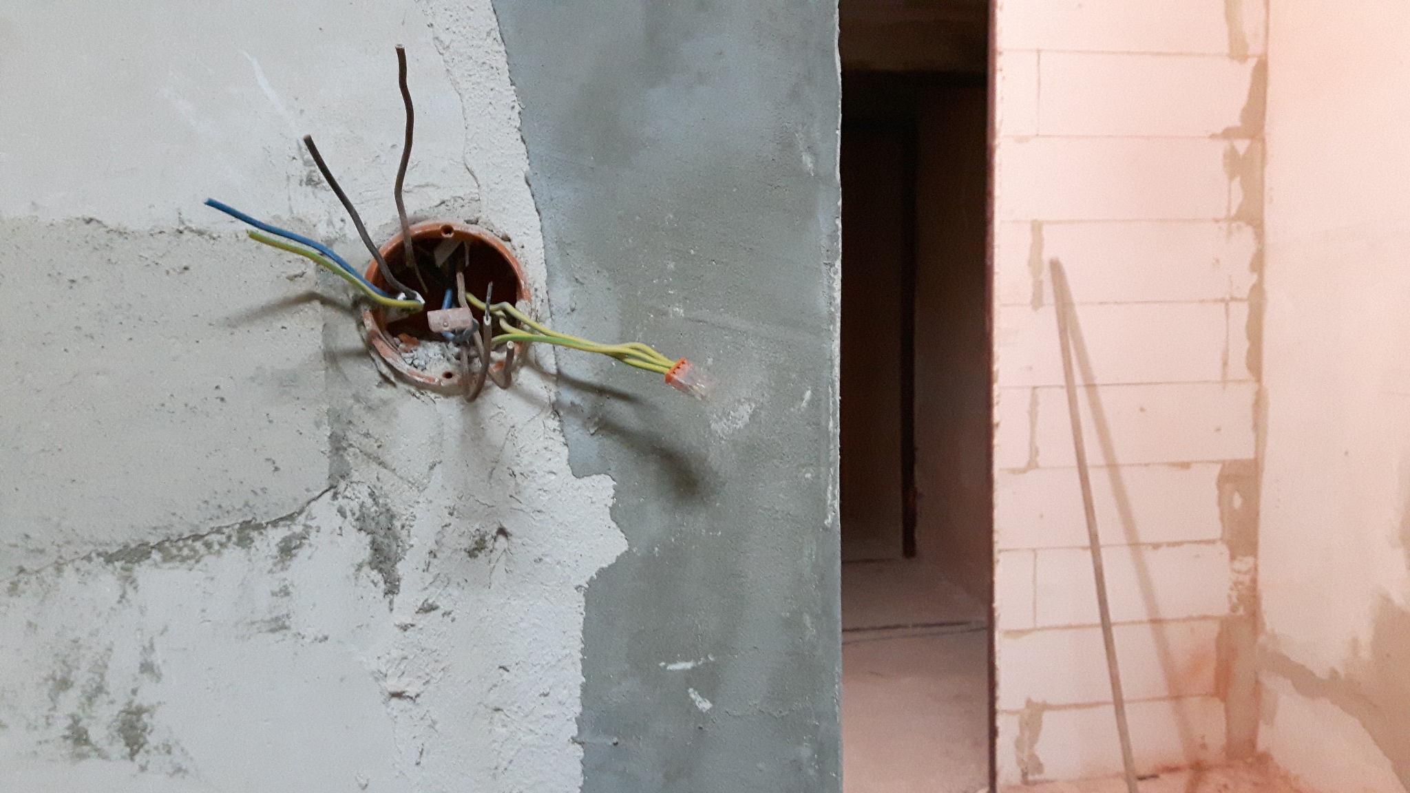 Niefrezowana puszka elektryczna z wystającymi przewodami na surowej ścianie, widoczne złącza WAGO. W tle fragment ściany z betonu komórkowego i otwór drzwiowy.