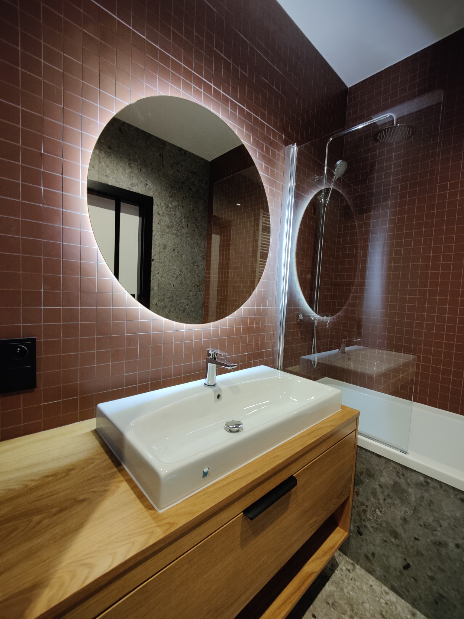 Nowoczesna łazienka z okrągłym lustrem LED, umywalką nablatową na drewnianej szafce i kabiną prysznicową. Brązowe płytki na ścianach i szary wzór lastryko na podłodze.