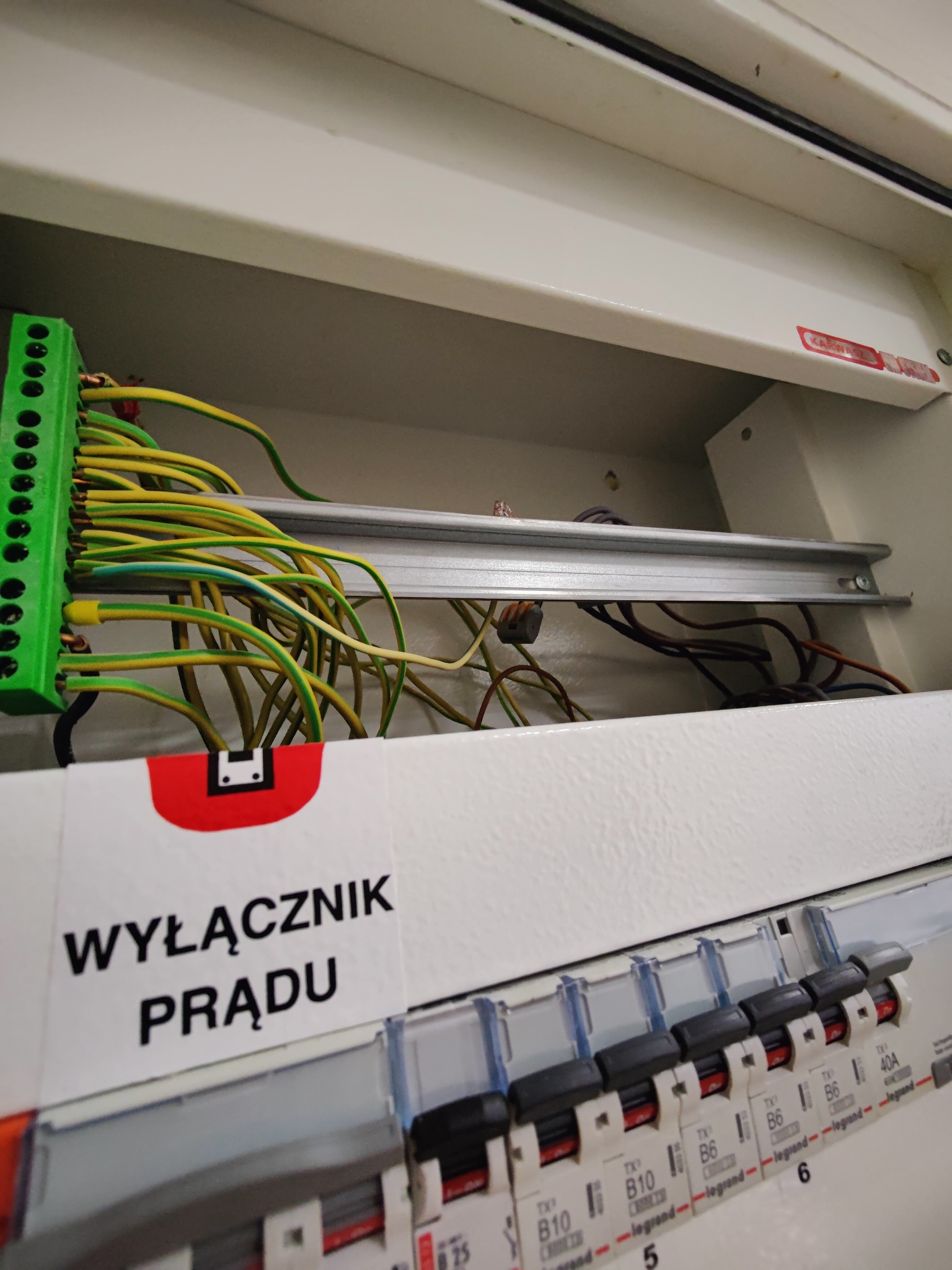 Otwarta skrzynka elektryczna z podłączonymi przewodami (żółto-zielone) do listwy zaciskowej. Widoczne bezpieczniki i etykieta 'Wyłącznik prądu'.