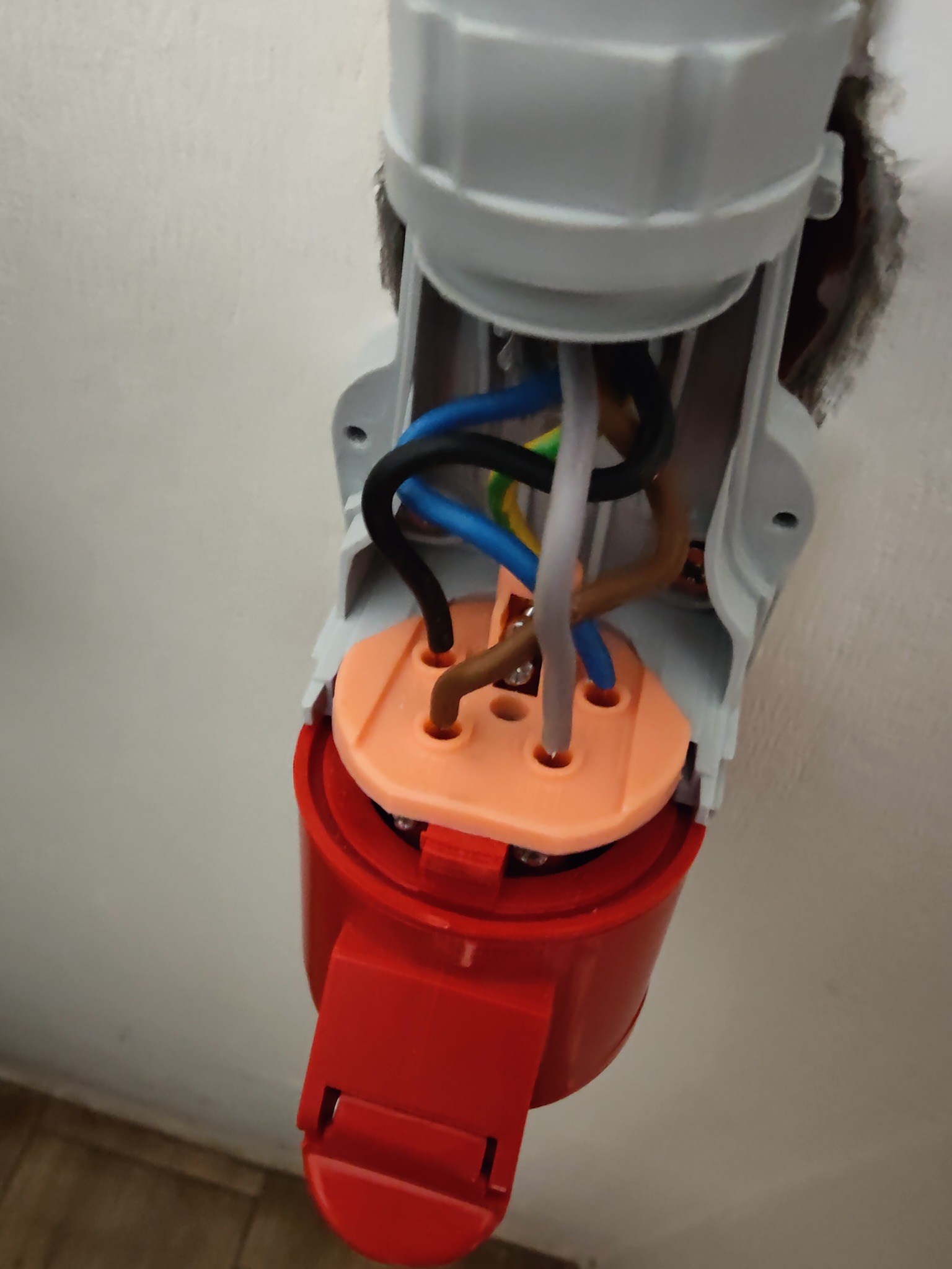 Otwarta, czerwono-szara wtyczka przemysłowa z widocznymi przewodami (czarny, niebieski, brązowy, biały, żółto-zielony) wystającymi ze ściany. Widok z bliska.