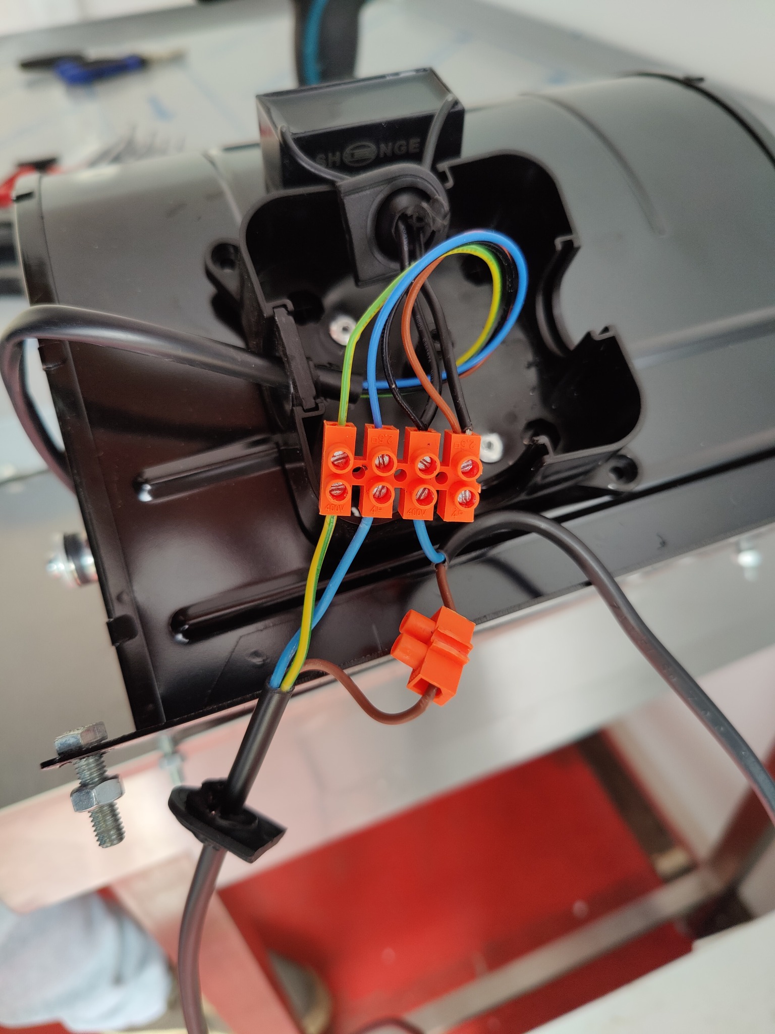 Czarny element z podłączonymi przewodami i pomarańczowymi złączkami. Widoczne śruby mocujące. Ujęcie z bliska, detale połączeń elektrycznych.