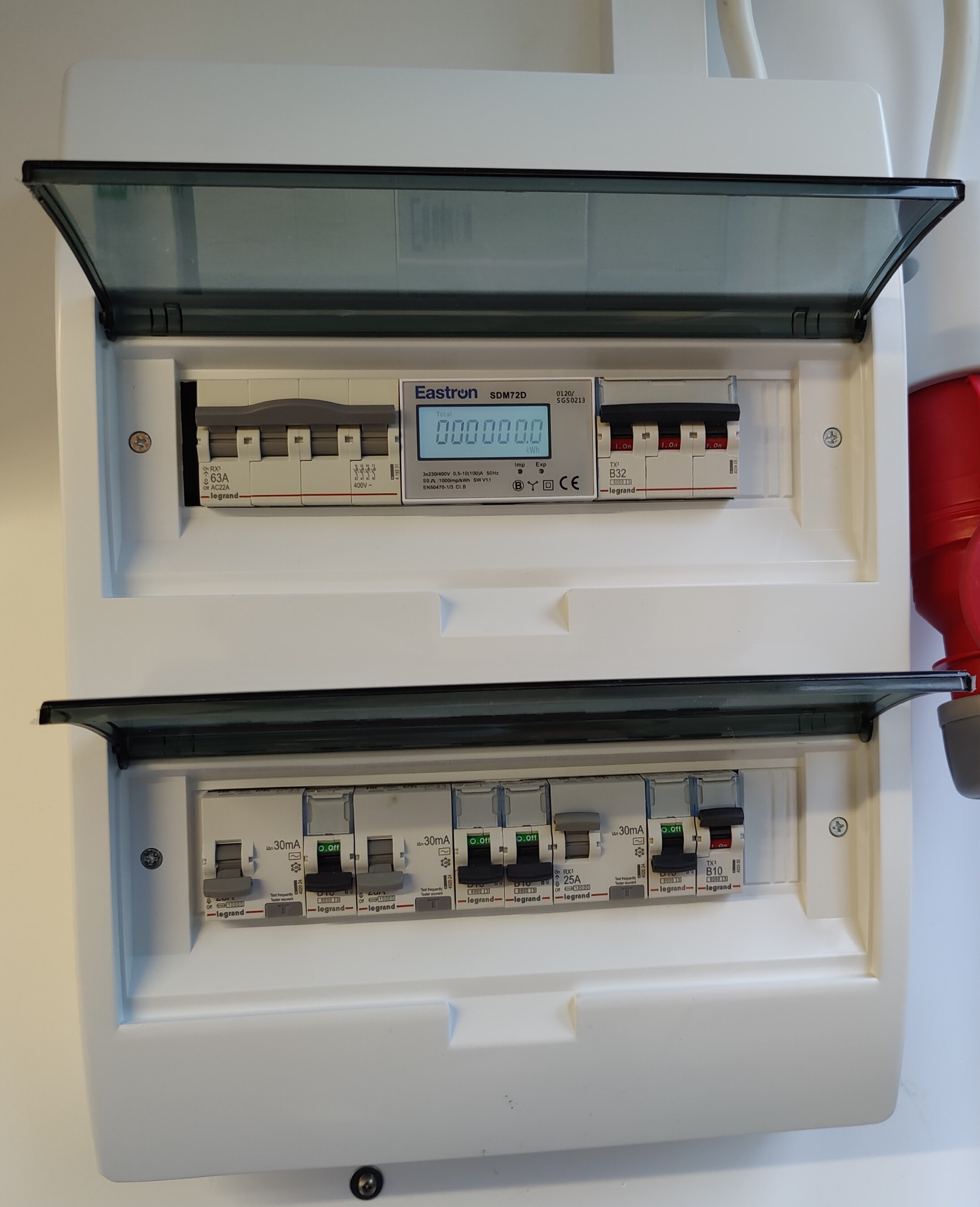 Rozdzielnica elektryczna z otwartymi klapami, widoczne zabezpieczenia i licznik energii. Biała obudowa, szare klapy, detale Legrand. Ujęcie z bliska, oświetlenie naturalne.