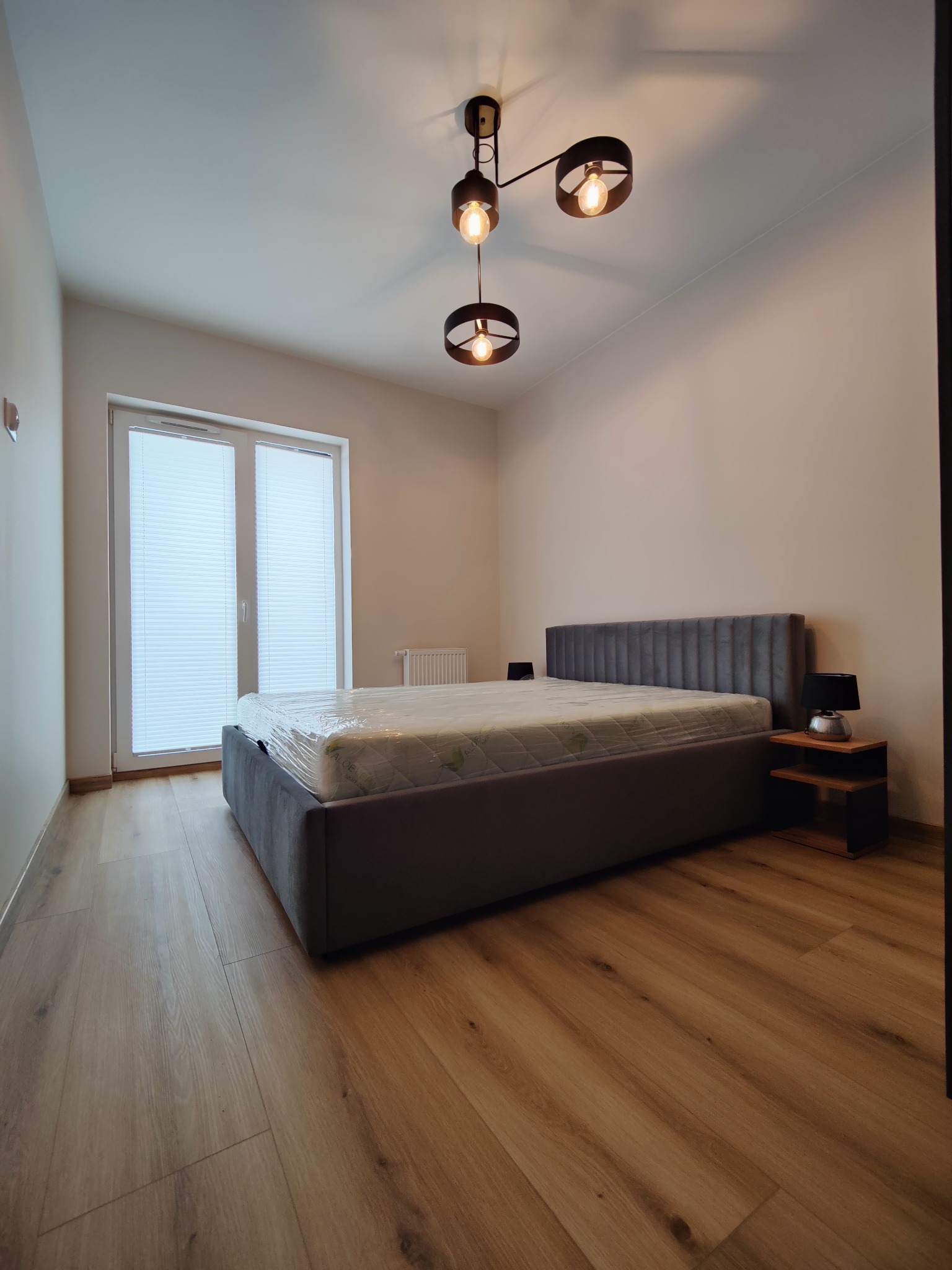 Nowoczesna sypialnia z szarym łóżkiem i drewnianą podłogą. Nad łóżkiem wisi designerska lampa z trzema żarówkami. Okno z żaluzjami w tle.