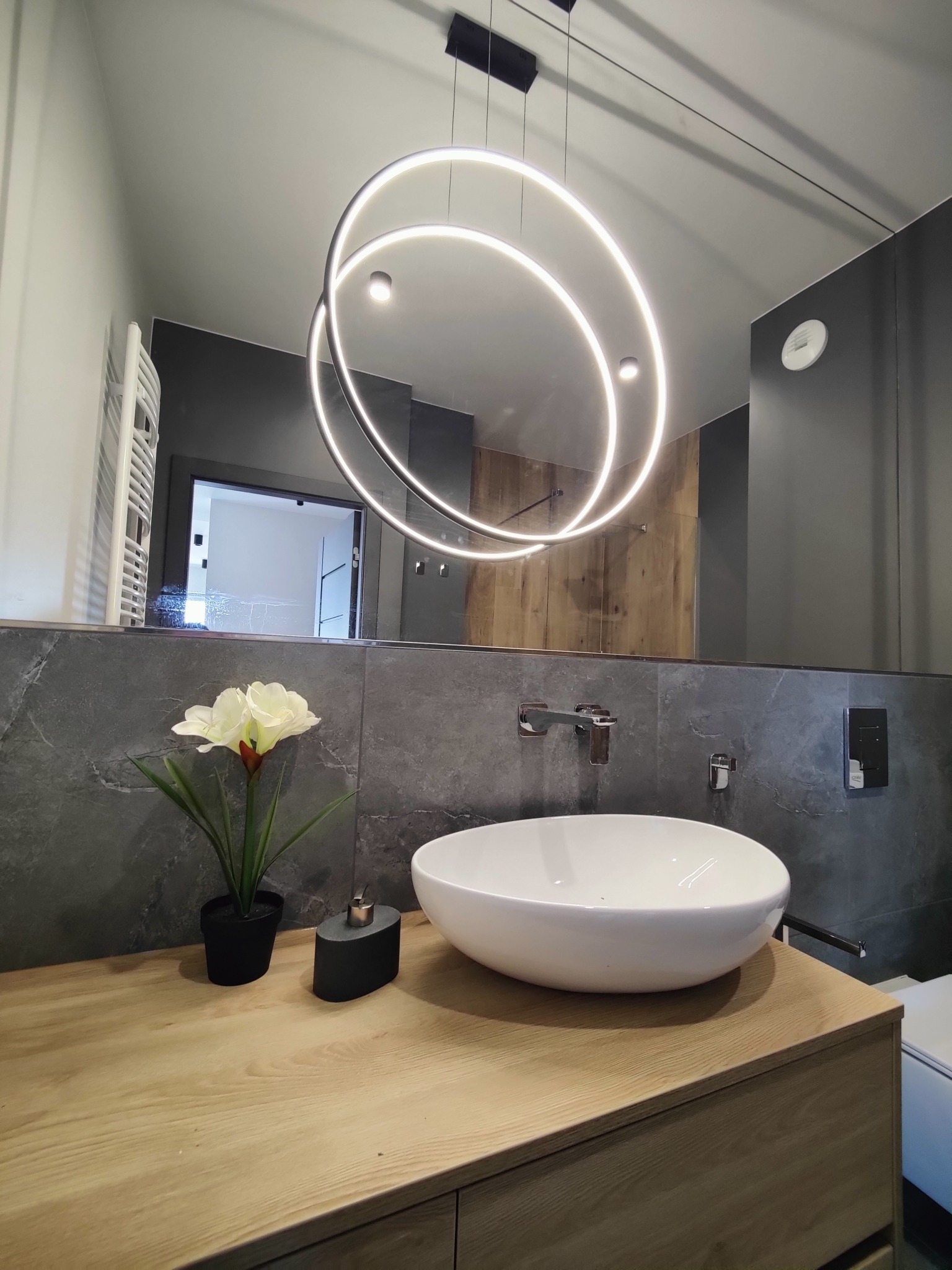 Nowoczesna łazienka z okrągłą lampą LED nad umywalką nablatową. Szare płytki i drewniany blat dodają elegancji. Widoczne detale armatury i dekoracje.