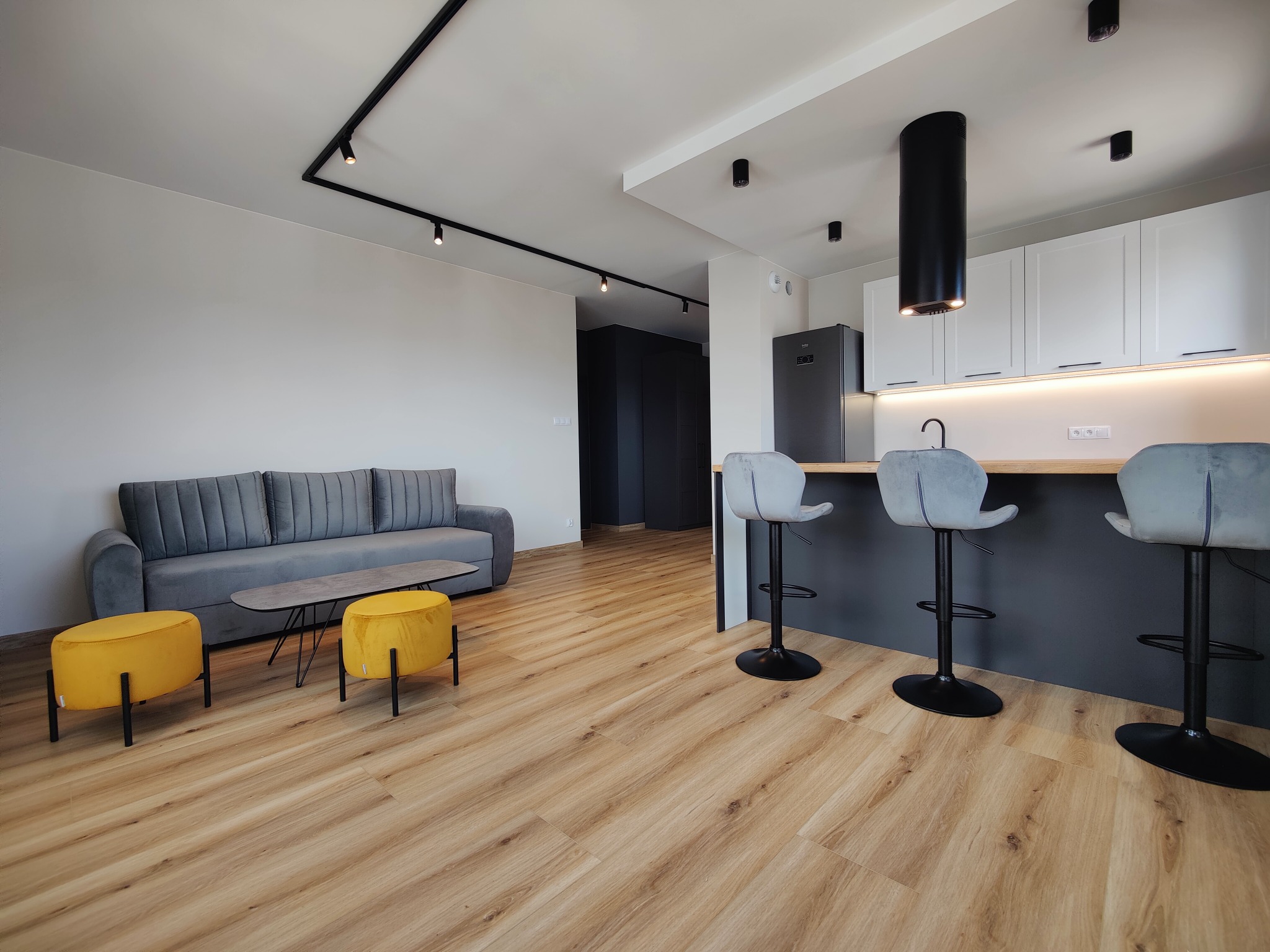 Nowoczesny salon z oświetleniem szynowym na suficie. Widoczne lampy punktowe, sofa, pufy i wyspa kuchenna z hokerami. Minimalistyczny design, jasne kolory i drewniana podłoga.