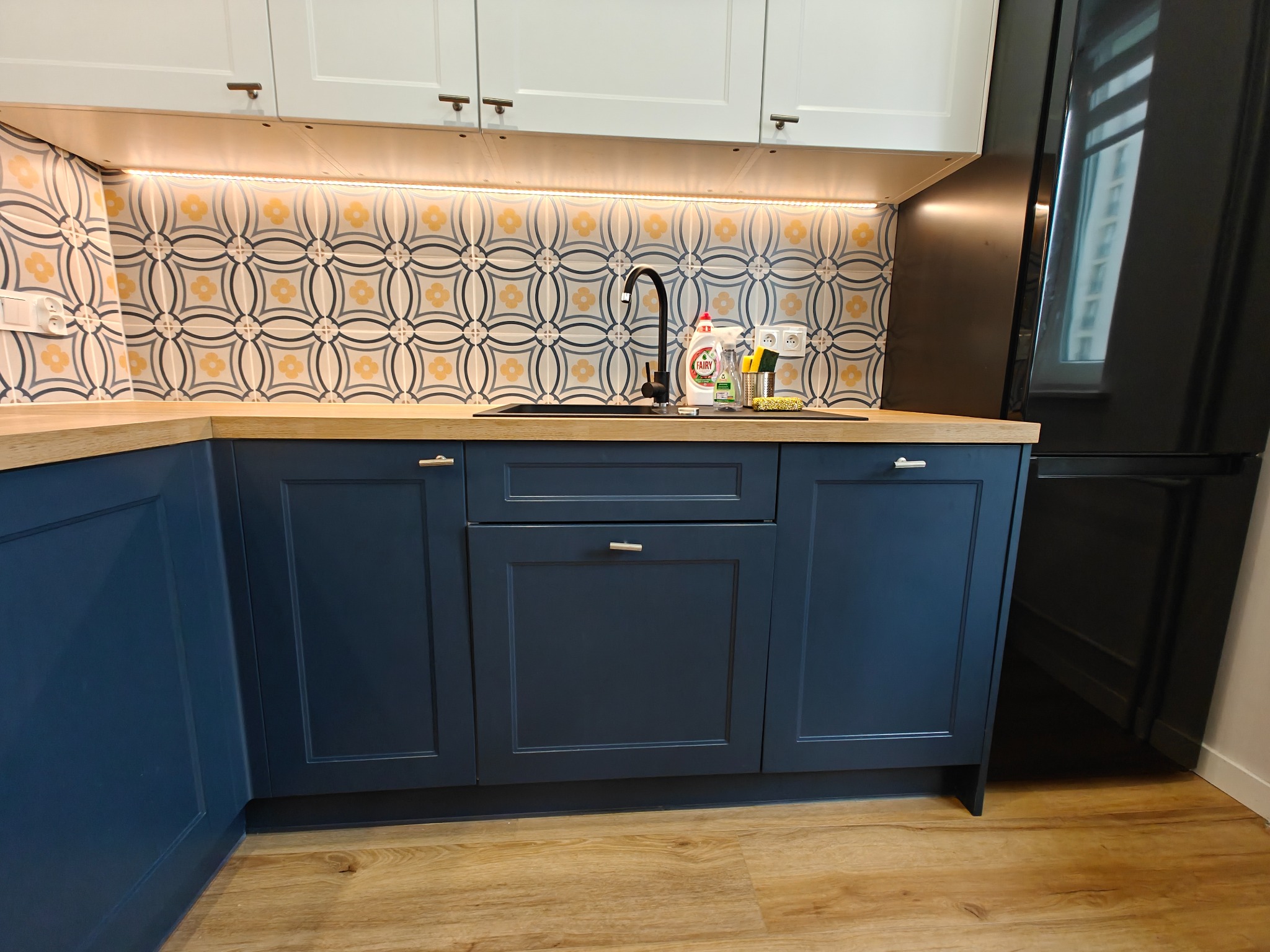 Niebieska zabudowa kuchenna z drewnianym blatem i wzorzystą mozaiką nad zlewem. Pod szafkami górnymi zamontowane oświetlenie LED.