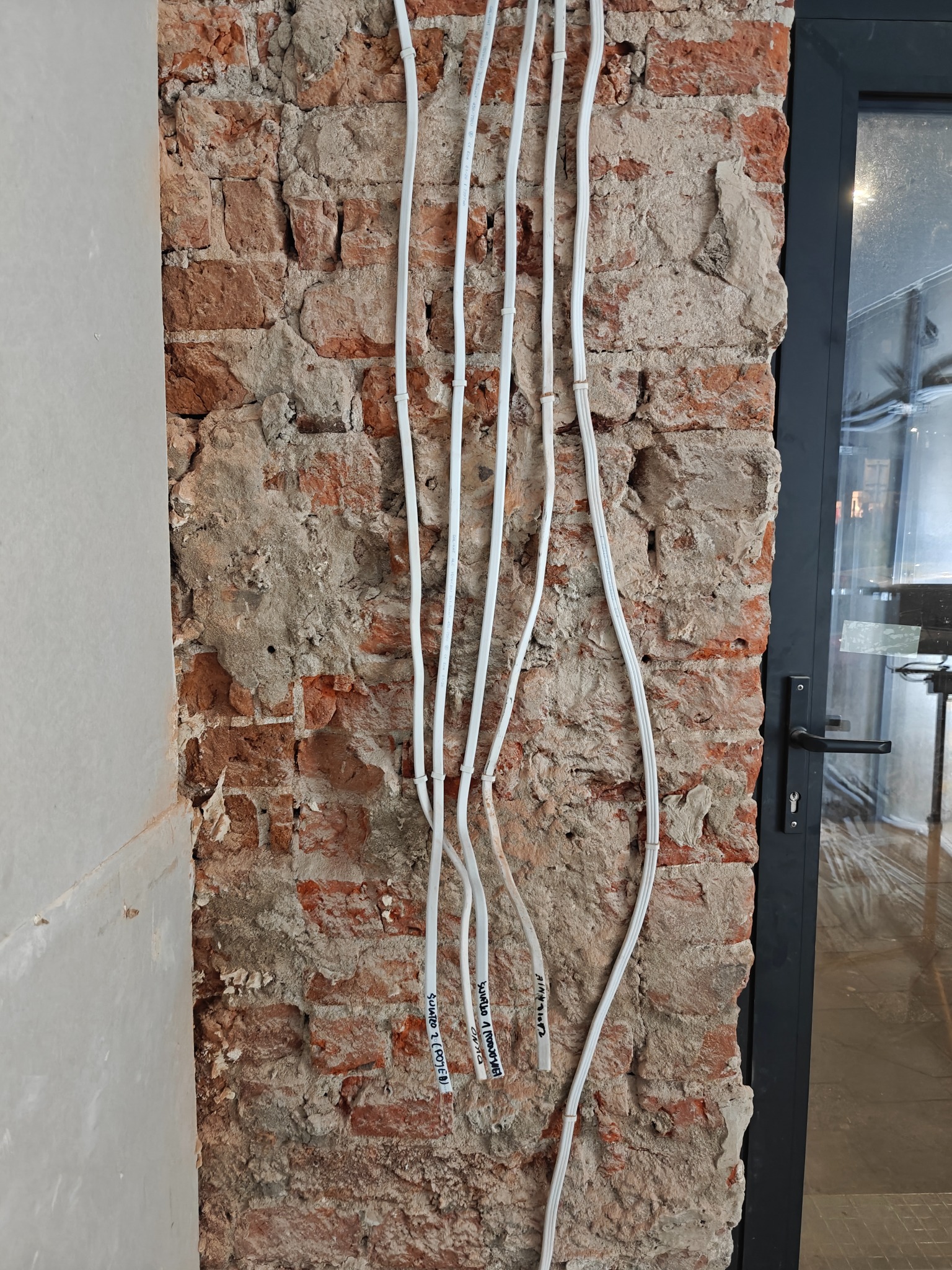 Białe przewody elektryczne prowadzone po surowej, ceglanej ścianie z widocznymi oznaczeniami, obok ściany otynkowanej i czarnej ramy okiennej.