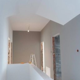 Ekspresowe Metamorfozy - Wnętrze po malowaniu na szaro, widoczna drabina malarska, surowe ościeżnice drzwiowe i fragment balustrady. Oświetlenie punktowe.