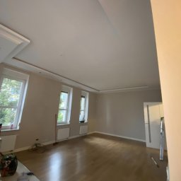 Ekspresowe Metamorfozy - Jasne, przestronne wnętrze po malowaniu: parkiet, ściany w odcieniach beżu, trzy okna z widokiem na zieleń. Minimalistyczny wystrój, listwy przysufitowe.