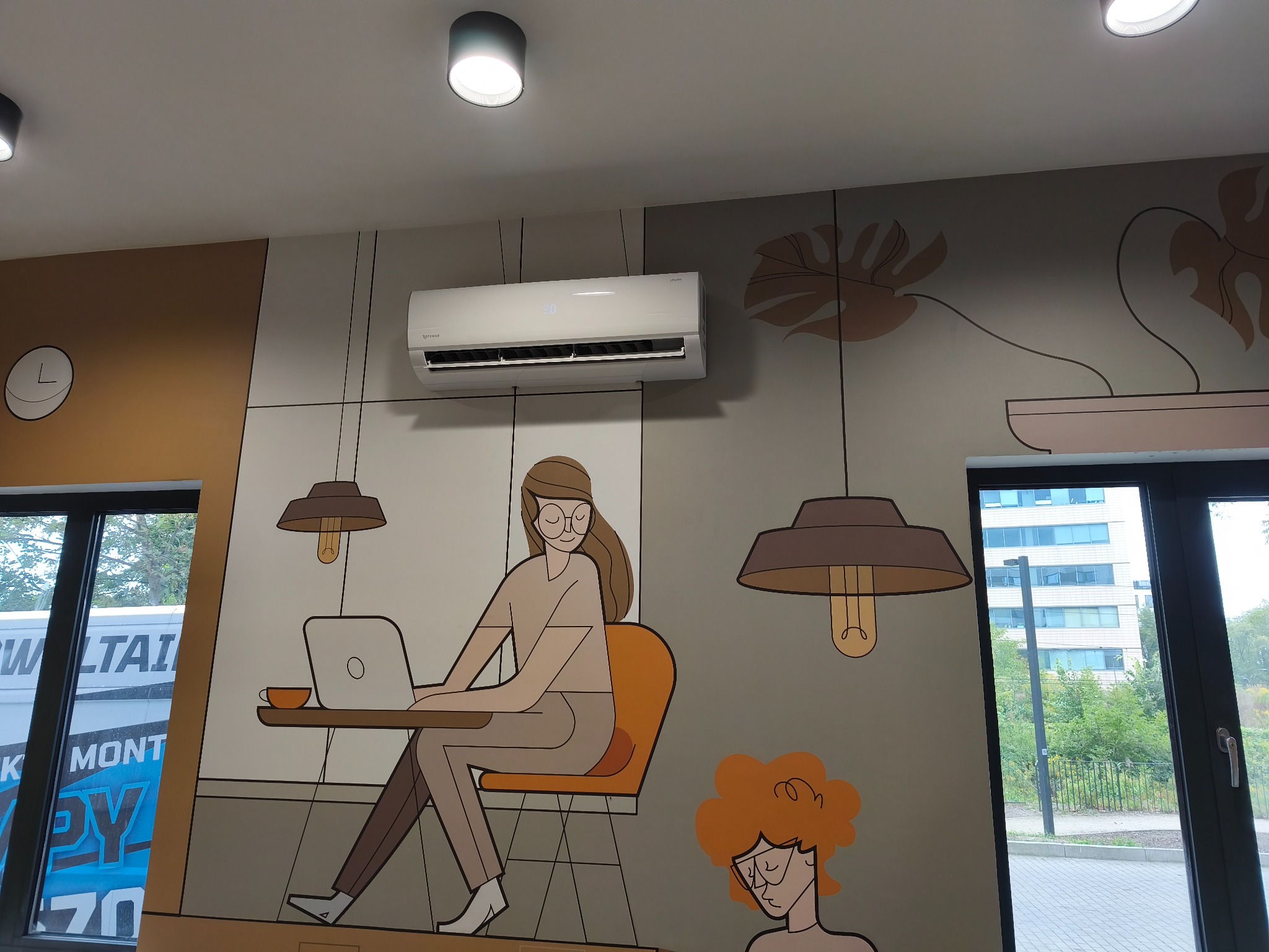 Biały klimatyzator na ścianie z rysunkami w biurze. Widoczne okno i lampy sufitowe. Nowoczesny design wnętrza z elementami graficznymi.