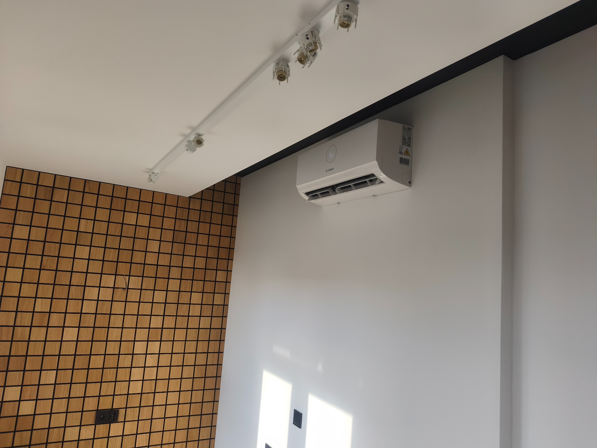 Biały klimatyzator Bosch zamontowany na ścianie w nowoczesnym wnętrzu z drewnianą mozaiką i oświetleniem szynowym.