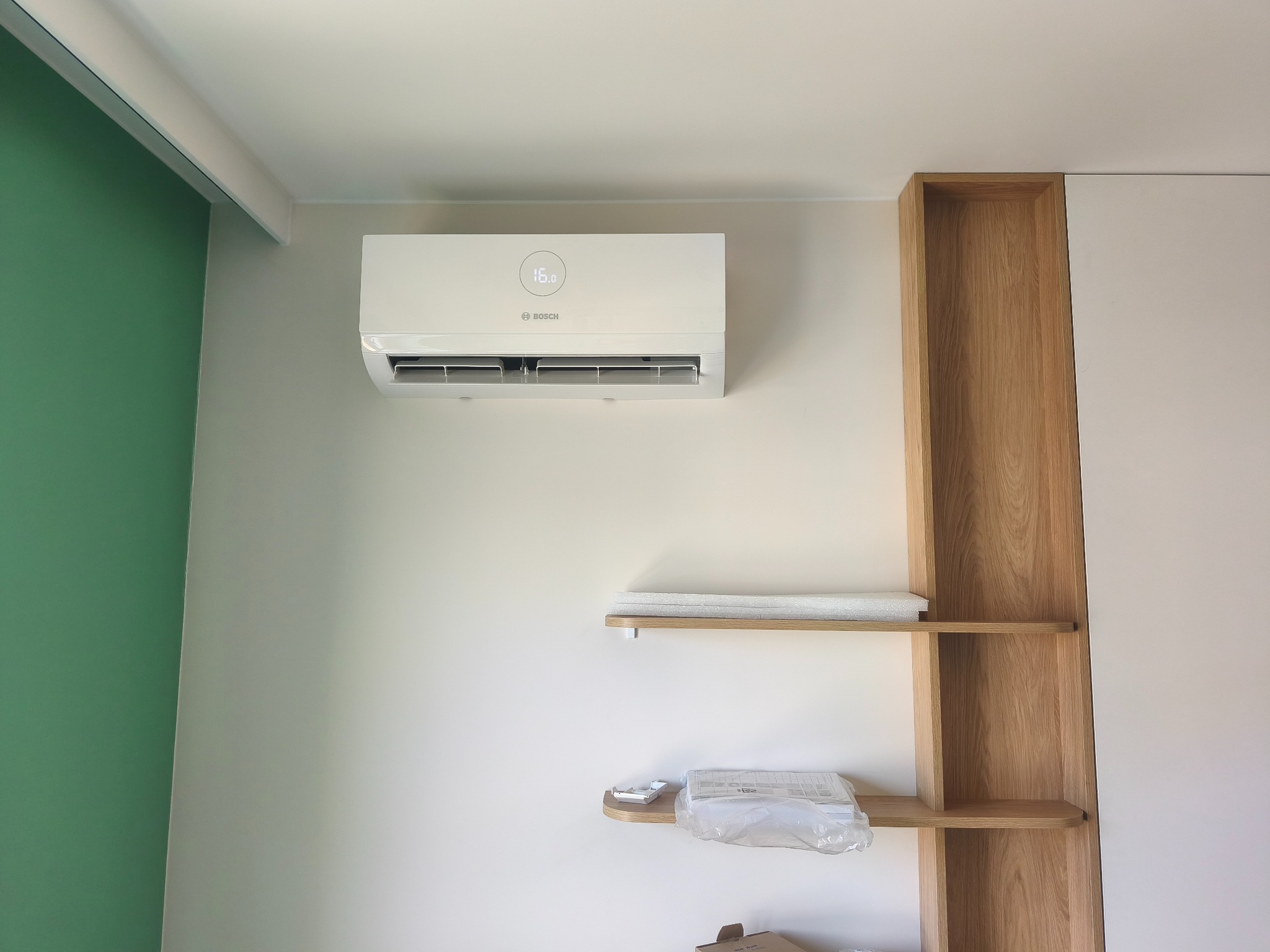 Biała klimatyzacja Bosch na ścianie w nowoczesnym wnętrzu z zielonym akcentem kolorystycznym i drewnianymi półkami. Widoczny wyświetlacz temperatury.