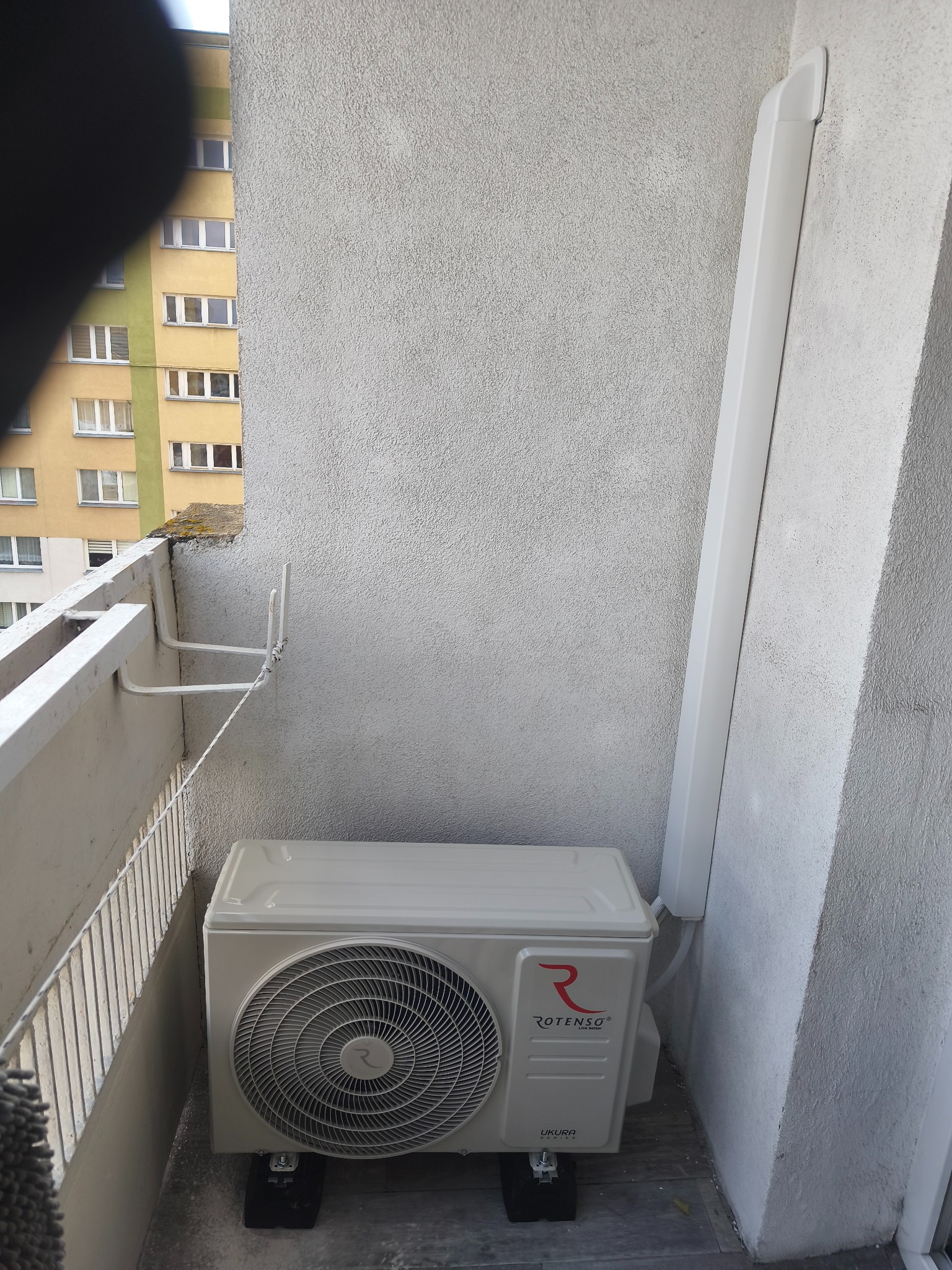 Biały klimatyzator Rotenso Ukura zamontowany na balkonie bloku mieszkalnego, z widoczną instalacją odprowadzającą skropliny. Szara ściana i fragment budynku w tle.
