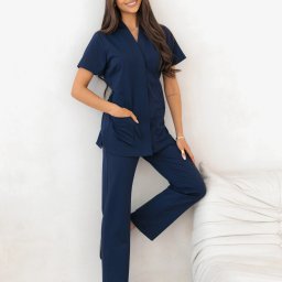 Veronica Medina - Uśmiechnięta brunetka w granatowym komplecie medycznym (bluza z krótkim rękawem i spodnie), oparta o białą ścianę i sofę. Widoczne czarne buty sportowe.