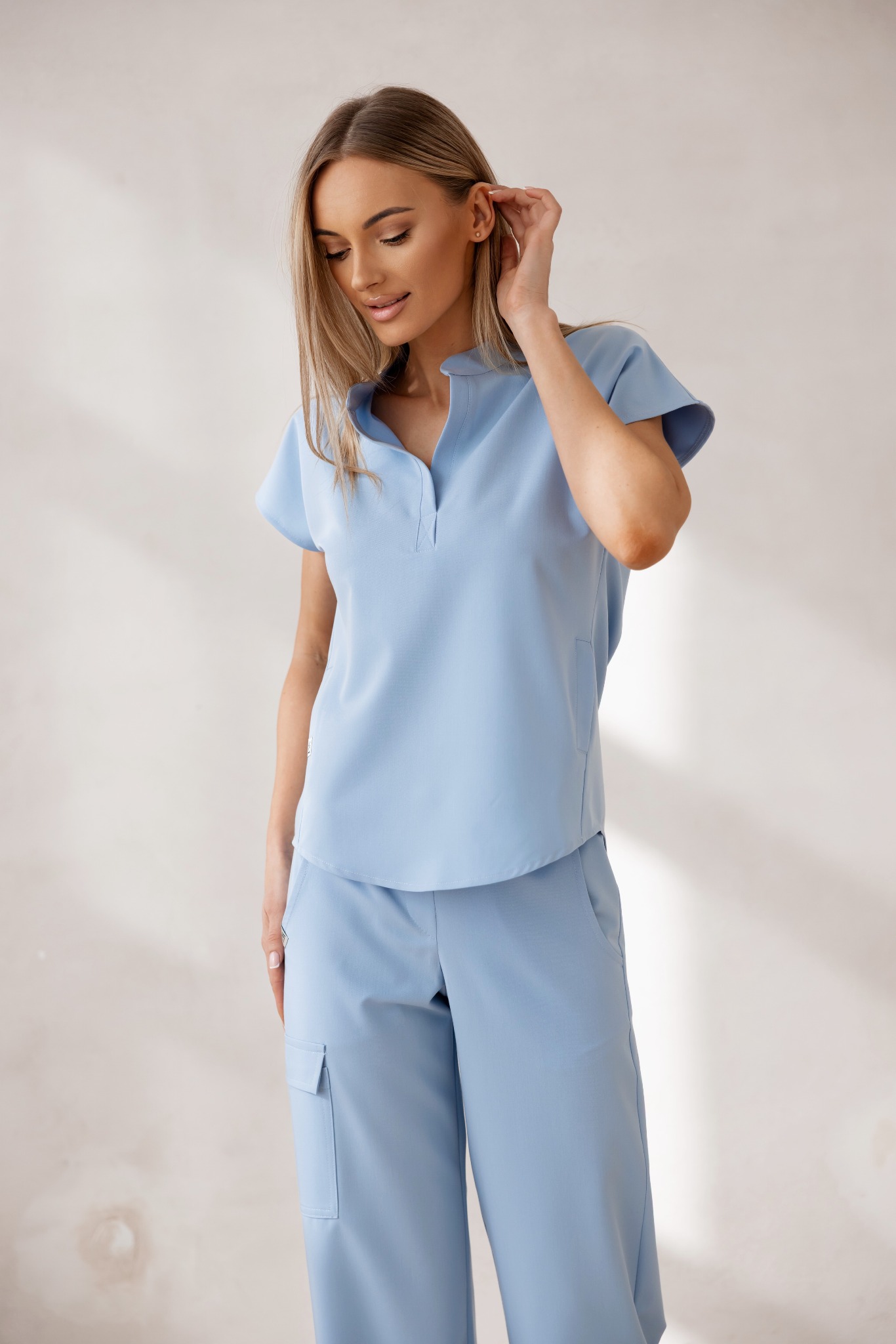 Kobieta w błękitnym komplecie: bluzka z krótkim rękawem i spodnie cargo. Modelka delikatnie dotyka włosów, spoglądając w dół. Stonowane tło, miękkie światło.