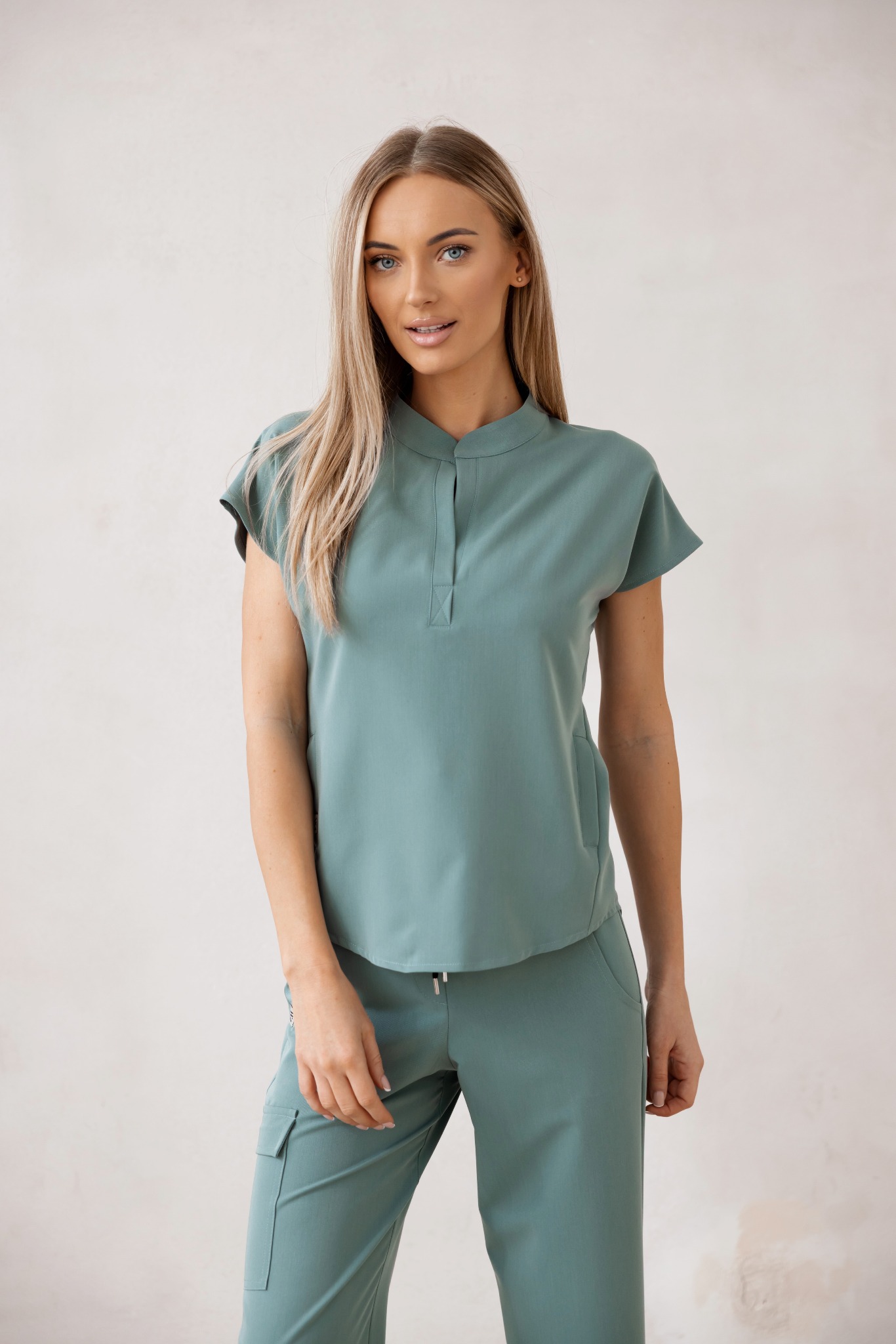 Uśmiechnięta blondynka w turkusowym komplecie: bluzka z krótkim rękawem i spodnie cargo. Stonowane tło, naturalne światło, pełny kadr.