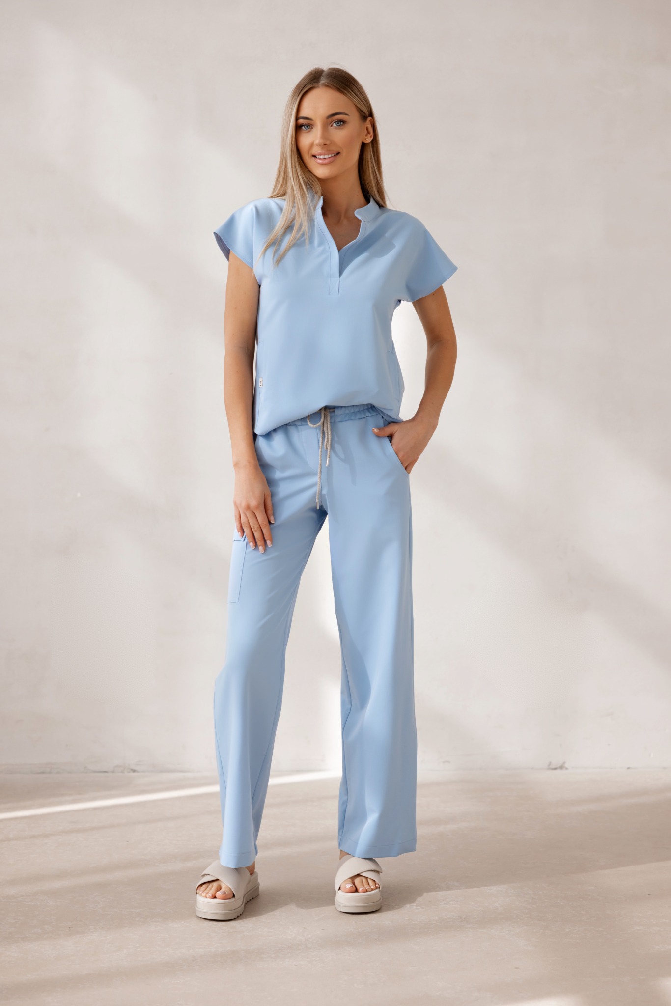 Kobieta prezentuje komplet odzieży medycznej w odcieniu błękitu, składający się z bluzki z krótkim rękawem i szerokich spodni. Całość w minimalistycznym stylu na jasnym tle.