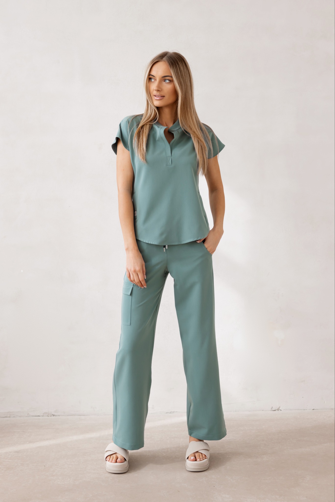 Kobieta prezentuje komplet odzieży medycznej w odcieniu morskiej zieleni. Bluza z krótkim rękawem i spodnie z kieszenią cargo. Całość na tle jasnej ściany.