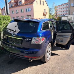 Piotr Kozłowiecki - Niebieska Mazda CX-7 z otwartymi drzwiami na tle miejskiego krajobrazu z budynkami i przystankiem autobusowym. Widok z tyłu, lekko z boku.