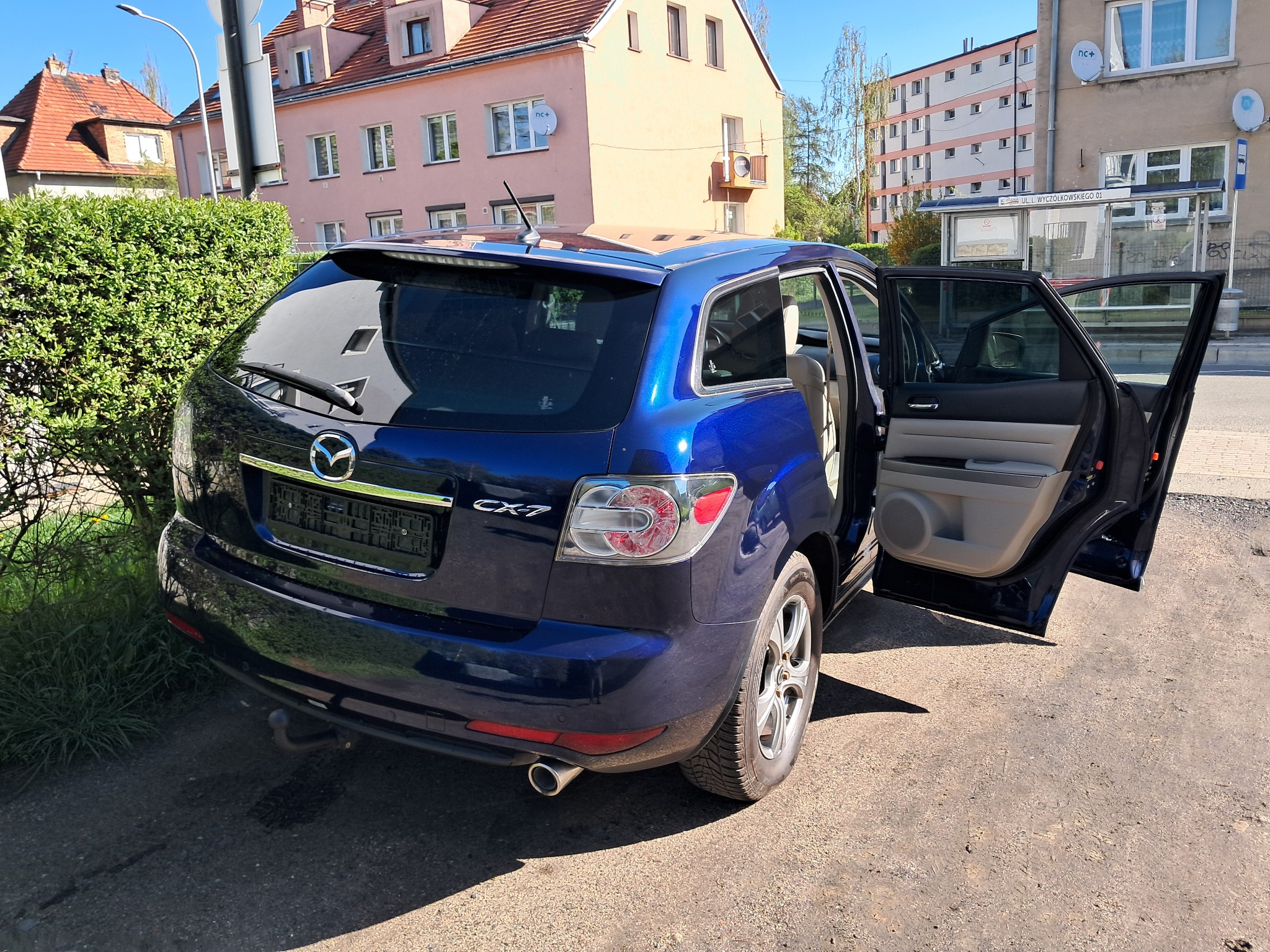 Niebieska Mazda CX-7 z otwartymi drzwiami na tle miejskiego krajobrazu z budynkami i przystankiem autobusowym. Widok z tyłu, lekko z boku.