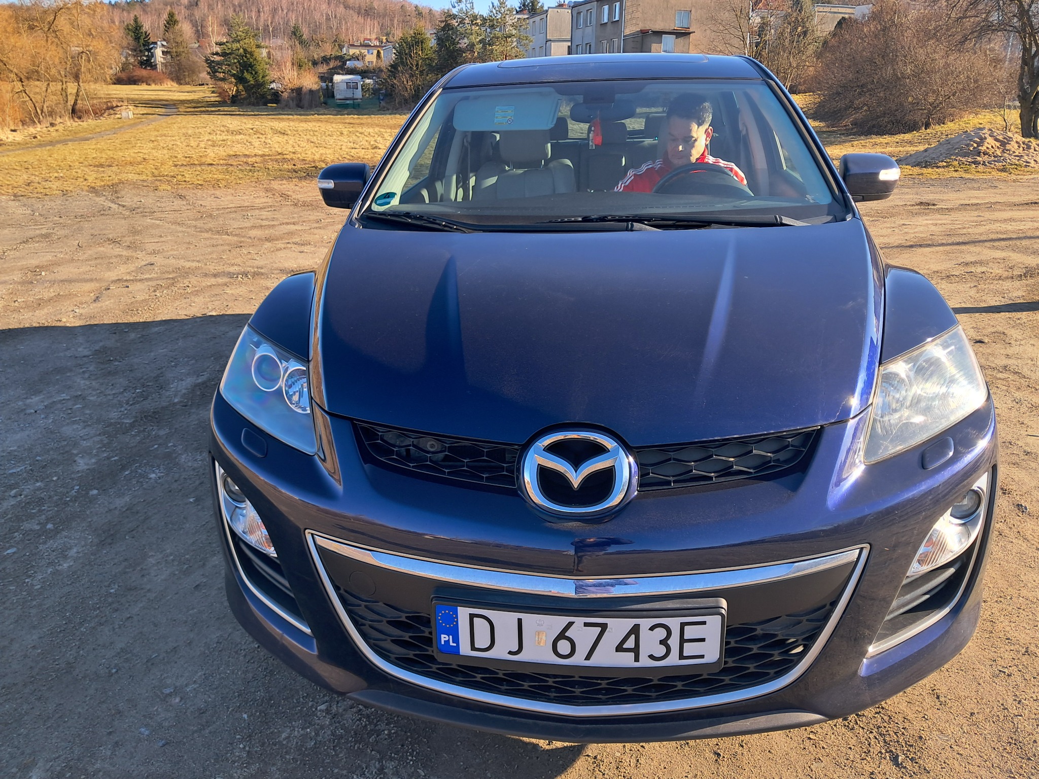 Niebieska Mazda CX-7 z kierowcą za kierownicą, zaparkowana na żwirowym podłożu w słoneczny dzień. Widoczny numer rejestracyjny DJ 6743E.