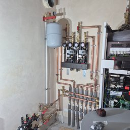 Dawid Sulej - Złożona instalacja grzewcza z miedzianymi rurami, manometrami i zbiornikiem wyrównawczym na tle surowej ściany. Widoczne elementy kotłowni i narzędzia.