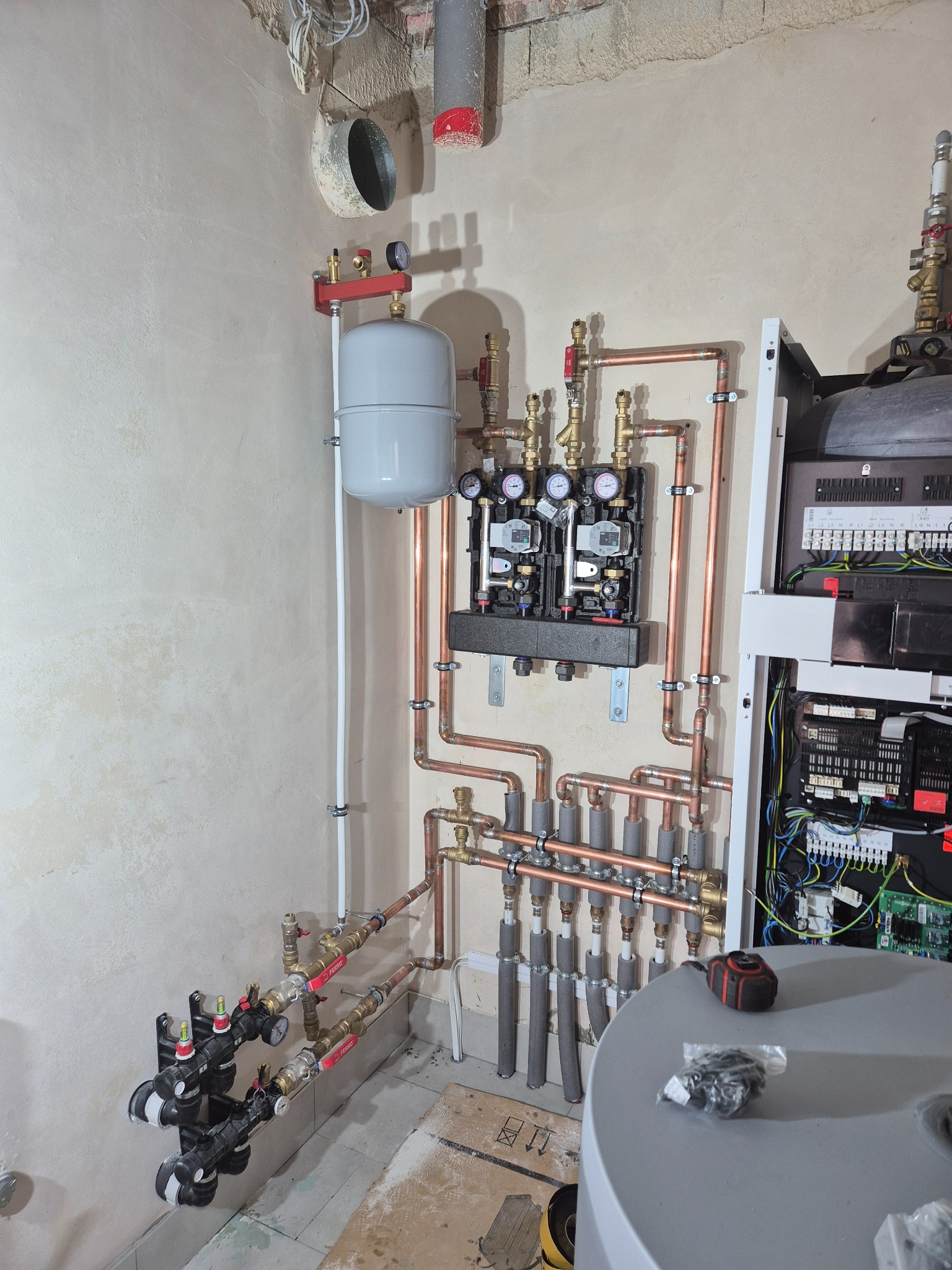 Złożona instalacja grzewcza z miedzianymi rurami, manometrami i zbiornikiem wyrównawczym na tle surowej ściany. Widoczne elementy kotłowni i narzędzia.