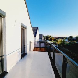 TOSIEZATEGUJE.PL SPÓŁKA Z OGRANICZONĄ ODPOWIEDZIALNOŚCIĄ - Biały balkon z przeszkloną balustradą w nowoczesnym domu, widok na okolicę w słoneczny dzień. Minimalistyczny design, czyste linie, jasne kolory.