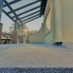 TOSIEZATEGUJE.PL SPÓŁKA Z OGRANICZONĄ ODPOWIEDZIALNOŚCIĄ - Wykończony taras z żywicy w Krakowie, zadaszony pergolą i roletami zewnętrznymi. Widok z niskiej perspektywy podkreśla strukturę powierzchni i detale wykończenia elewacji.