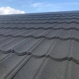 TOSIEZATEGUJE.PL SPÓŁKA Z OGRANICZONĄ ODPOWIEDZIALNOŚCIĄ - Szczegółowe ujęcie nowo położonego, grafitowego dachu z blachodachówki modułowej o falistym wzorze, na tle błękitnego nieba z delikatnymi obłokami.