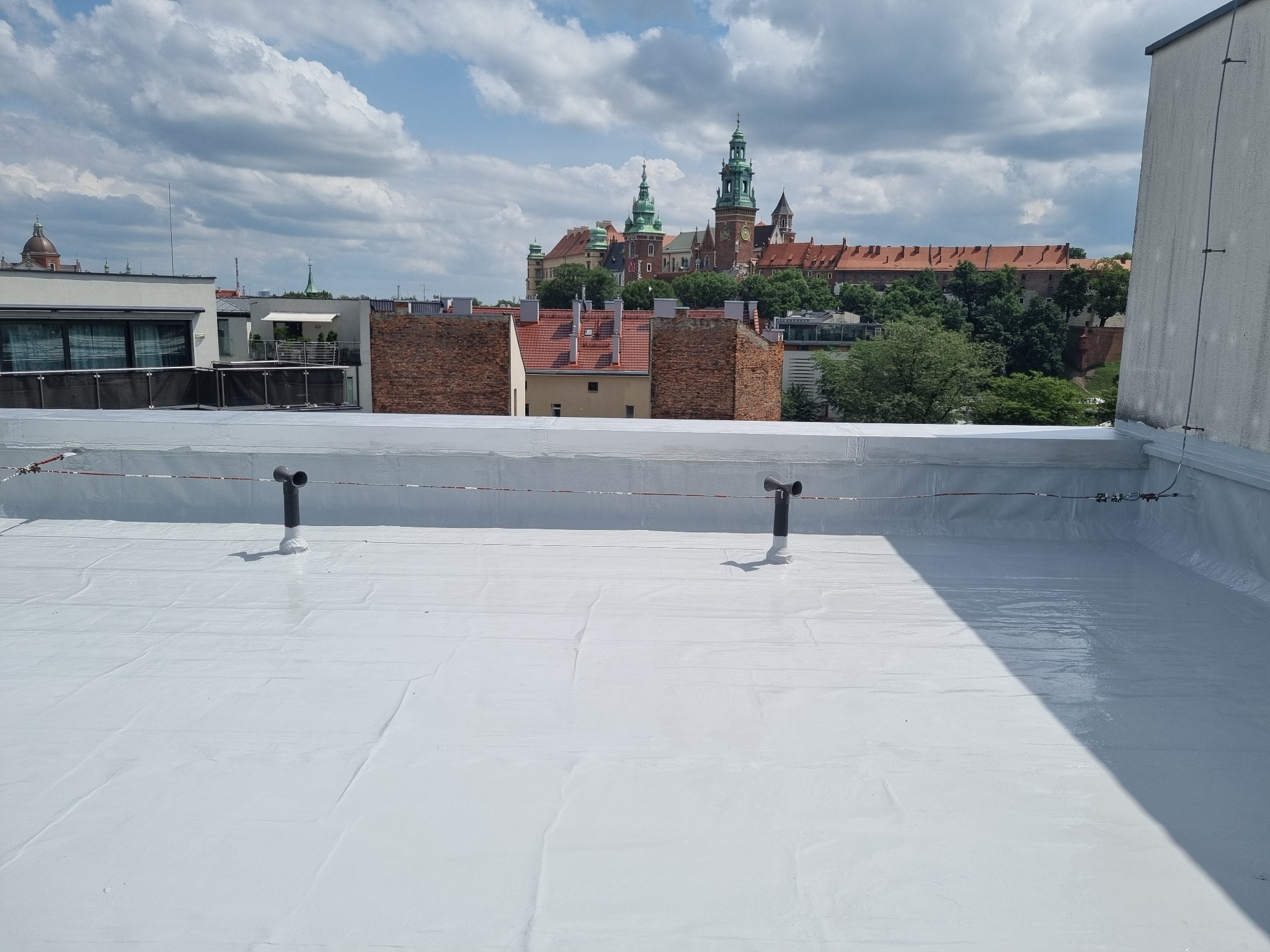 Biały, płaski dach po renowacji z dwoma pionowymi rurami wentylacyjnymi na tle panoramy miasta z Wawelem w Krakowie, widoczne łączenia papy.