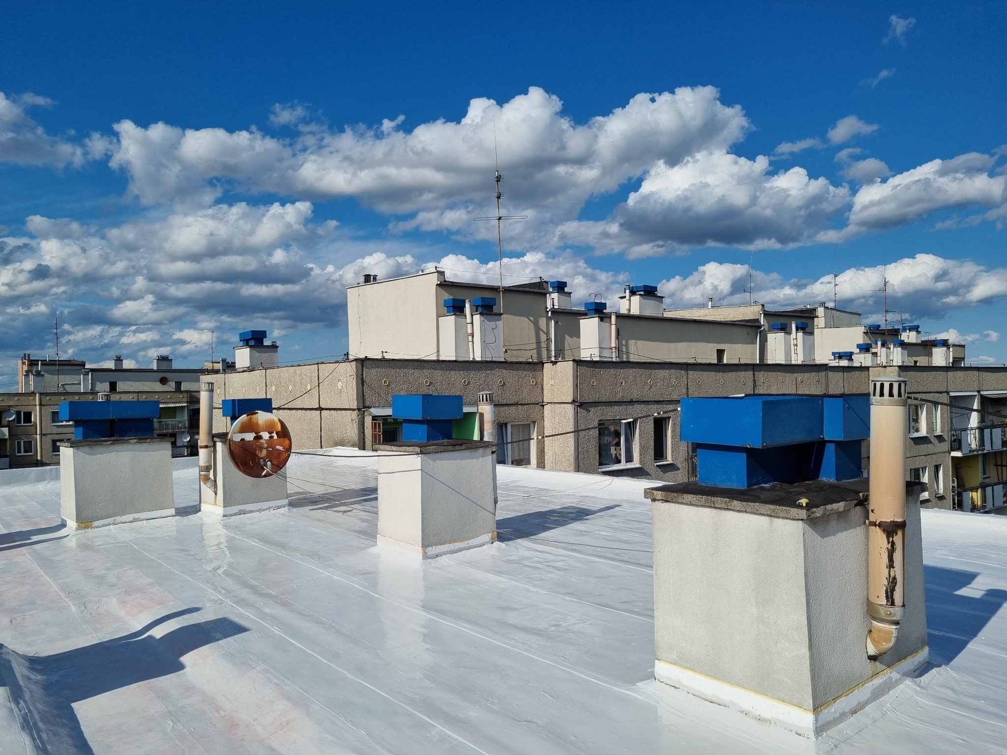 Biała, świeżo pokryta powłoką dachową powierzchnia z kominami na tle błękitnego nieba z chmurami. Widok na budynki mieszkalne w oddali.