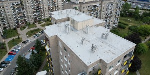 Jasnoszary, płaski dach bloku z kominami wentylacyjnymi, widziany z góry. W tle inne budynki i zieleń. Ujęcie z drona, lekko z prawej strony.