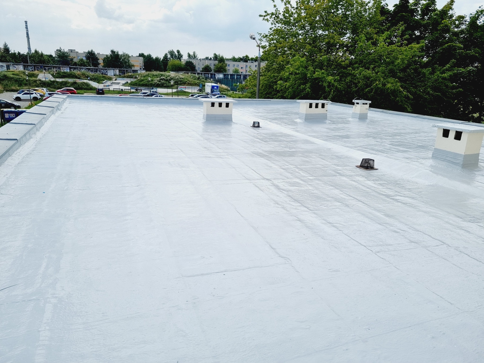 Jasnoszary, płaski dach budynku po renowacji z kominami wentylacyjnymi. W tle zieleń drzew i zabudowa miejska. Widok z góry, szeroki kadr.