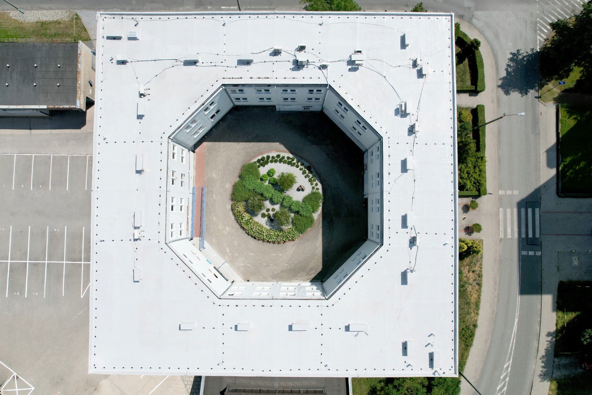 Widok z drona na biały, ośmiokątny dach budynku z wewnętrznym dziedzińcem i okrągłym klombem. Widoczne elementy instalacji na dachu i fragment parkingu.