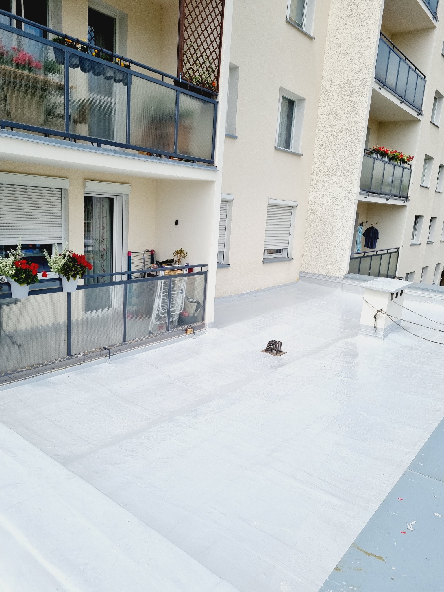 Biała, płaska powierzchnia dachu budynku wielorodzinnego z balkonami. Widoczny komin i kratka odpływowa. Balkony z kwiatami i meblami.