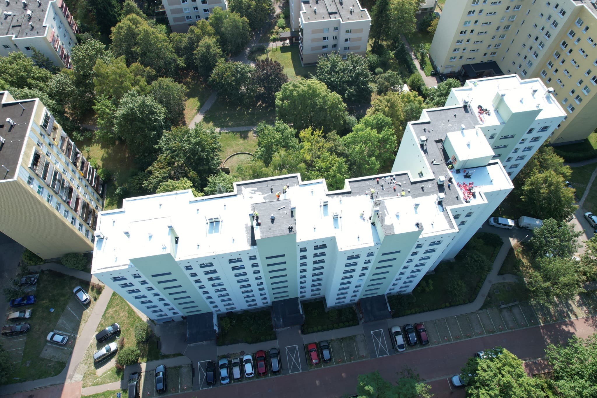 Z lotu ptaka: ekipa na jasnym dachu wysokiego bloku, prawdopodobnie podczas prac remontowych. Widoczne drzewa, parking i inne budynki w tle.