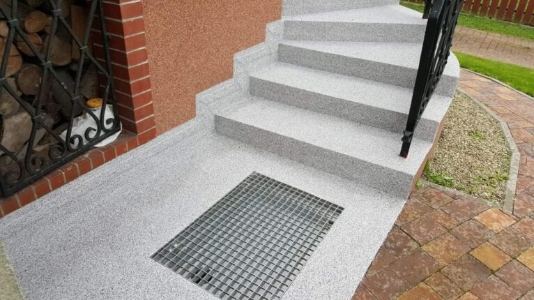 Schody zewnętrzne i podest wykończone szarą, żywiczną posadzką z drobnym kamyczkiem. Widoczna kratka ściekowa i fragment elewacji z cegły. Metalowa balustrada.