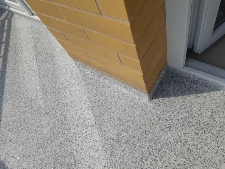 Szara, drobnoziarnista posadzka balkonu z ceglanym filarem i białym obramowaniem drzwi balkonowych. Widok z góry, oświetlenie dzienne.