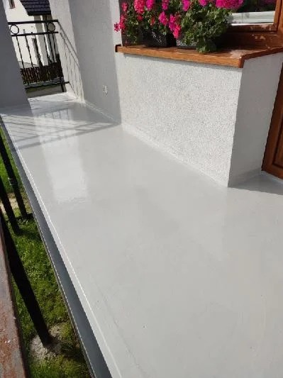 Balkon po renowacji z szarą, gładką powierzchnią, kwiatami w doniczce i widokiem na zieleń. Świeżo pomalowana podłoga balkonu.