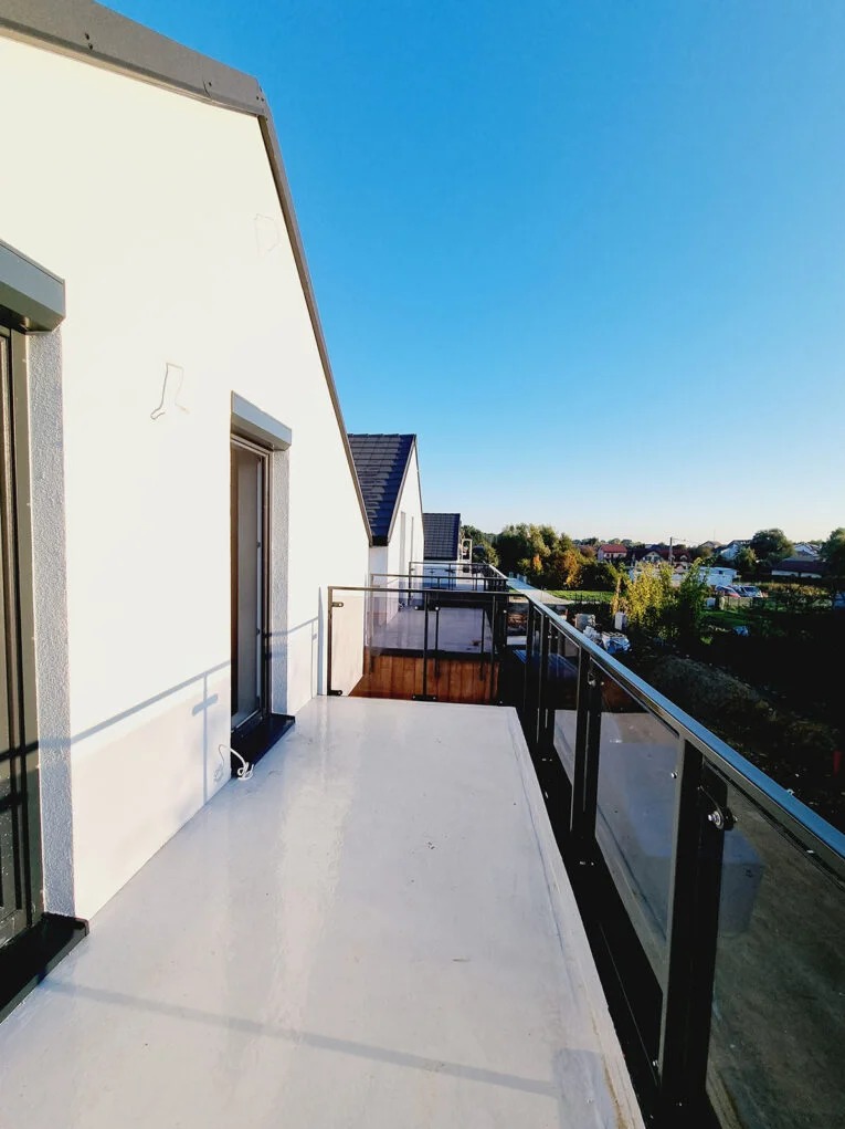 Biały balkon z przeszkloną balustradą w nowoczesnym domu, widok na okolicę w słoneczny dzień. Minimalistyczny design, czyste linie, jasne kolory.