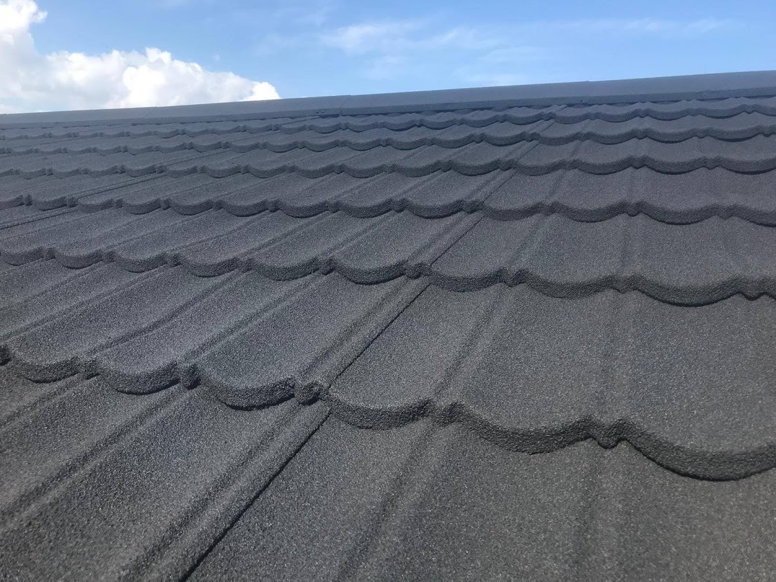 Szczegółowe ujęcie nowo położonego, grafitowego dachu z blachodachówki modułowej o falistym wzorze, na tle błękitnego nieba z delikatnymi obłokami.