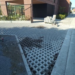 BRUK-OSZ - Układanie ażurowej kostki betonowej na podjeździe. Widoczny fragment niezasypanej nawierzchni, paleta kostki i zagęszczarka w tle nowoczesnego domu.