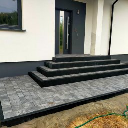 BRUK-OSZ - Nowoczesne schody wejściowe i chodnik z szarej kostki brukowej, obrzeża w kolorze antracyt. Widoczne przygotowane podłoże. Dom z białymi ścianami i szarą stolarką.