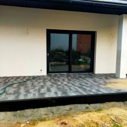 BRUK-OSZ - Nowoczesny taras z szarej kostki brukowej ułożonej przy domu z czarnymi ramami okiennymi. Widoczny fragment węża ogrodowego i niedokończone obrzeże.
