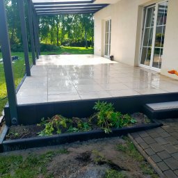 BRUK-OSZ - Nowoczesny taras z jasnych płyt, zadaszony pergolą. Widoczne elementy wykończeniowe, rabata z roślinami i fragment ogrodu. Świeże, poranne światło.