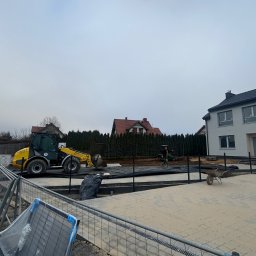 Firma Usługowo-Handlowa Andrzej Mydło - Żółta ładowarka teleskopowa na placu budowy z częściowo ułożoną kostką. W tle domy i pracownik z taczką. Widoczne tymczasowe ogrodzenie budowlane.