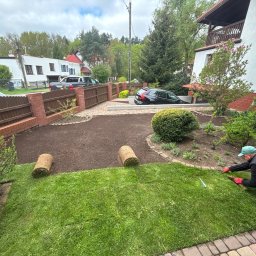 Firma Usługowo-Handlowa Andrzej Mydło - Układanie trawy z rolki w ogrodzie. Widoczny fragment trawnika, rolki trawy i osoba w trakcie pracy, w tle dom i ogrodzenie z cegły.